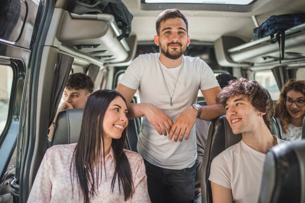 Portrait of a smiling young man standing in a bus next to a seat where his friends are sitting and looking directly at the camera